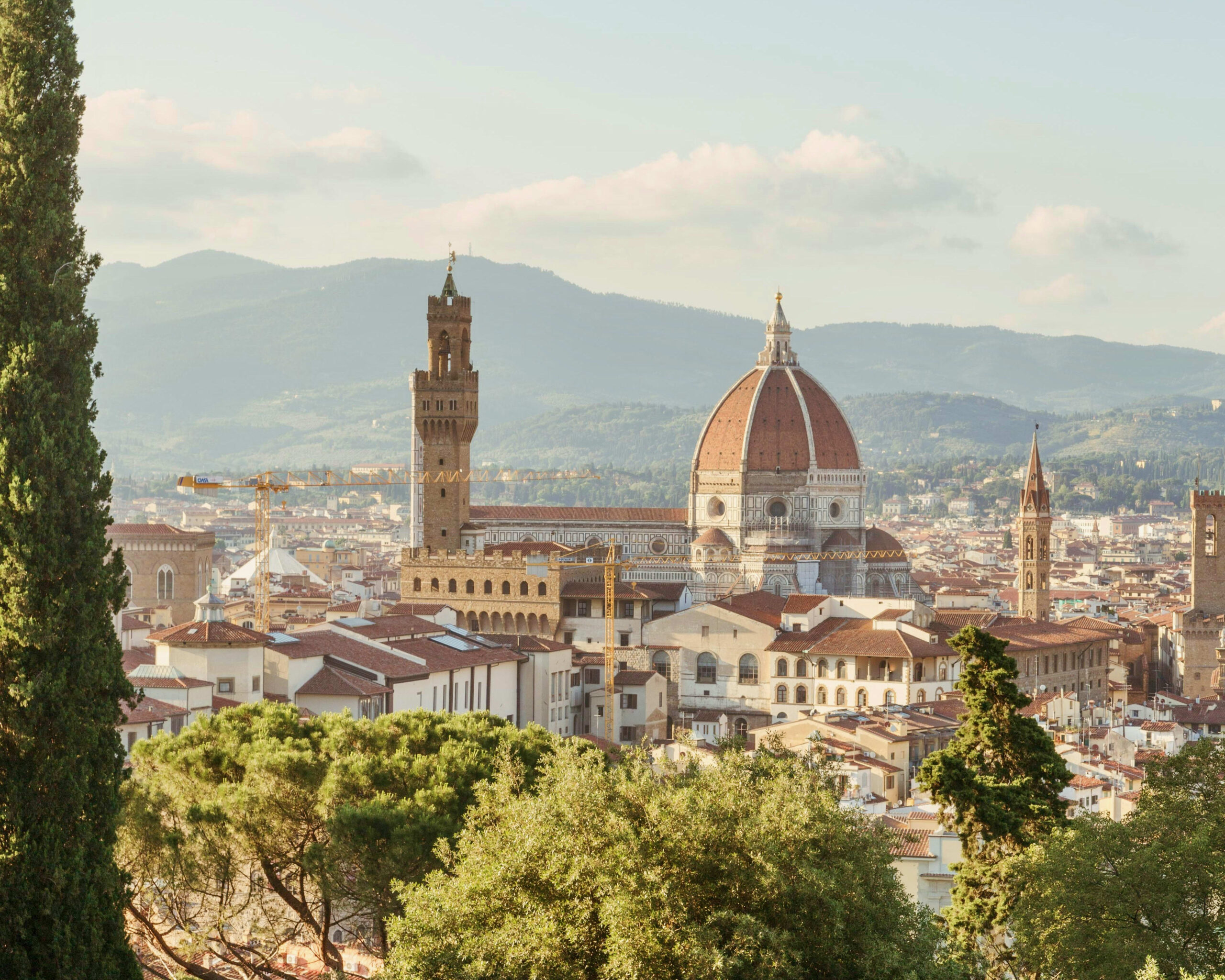 red rooftops across a city with a tower and red dome cathedral in the center, trees in the foreground, and mountains in the distance